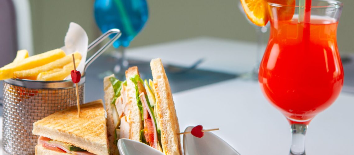 Fast food with sandwich, french fries, red cocktail, fork and knife on table, side view.